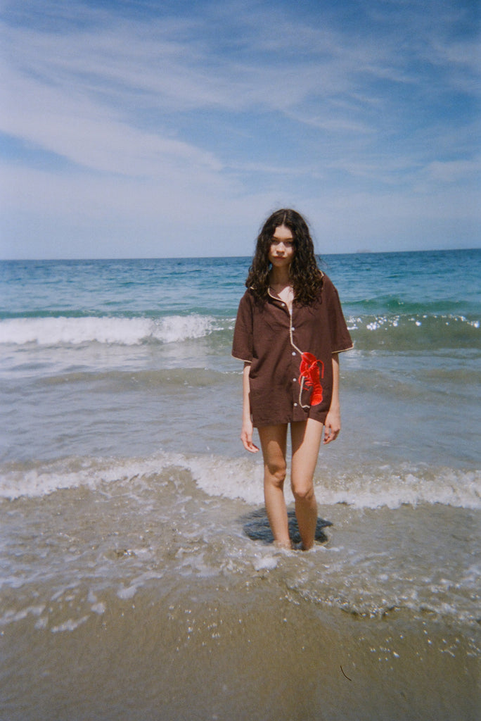 Female modeled pictured walking towards the camera at the beach, wearing the short-sleeved brown button-down ('Bisque Averse') over a swim suit. 