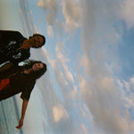 Sideways shot of two models standing ankle-deep in ocean water at sunset. The male model wears a black short-sleeve button-down shirt embroidered with two seahorses ('Tip your Bartenders' Cabana), paired with matching black trousers. The female model wears a brown short-sleeve shirt with beige piping and a large red lobster applique on the lower front ("Bisque Averse"), paired with white open-knit pants. They stand close together.