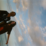 Sideways shot of two models standing ankle-deep in ocean water at sunset. The male model wears a black short-sleeve button-down shirt embroidered with two seahorses ('Tip your Bartenders' Cabana), paired with matching black trousers. The female model wears a brown short-sleeve shirt with beige piping and a large red lobster applique on the lower front ("Bisque Averse"), paired with white open-knit pants. They stand close together.