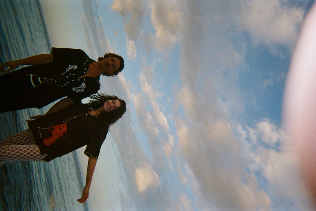 Sideways shot of two models standing ankle-deep in ocean water at sunset. The male model wears a black short-sleeve button-down shirt embroidered with two seahorses ('Tip your Bartenders' Cabana), paired with matching black trousers. The female model wears a brown short-sleeve shirt with beige piping and a large red lobster applique on the lower front ("Bisque Averse"), paired with white open-knit pants. They stand close together.