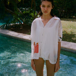Woman standing in a pool wearing a sheer white short-sleeve shirt with a red gingham pocket embroidered with French fries and a seagull embroidered on the sleeve.