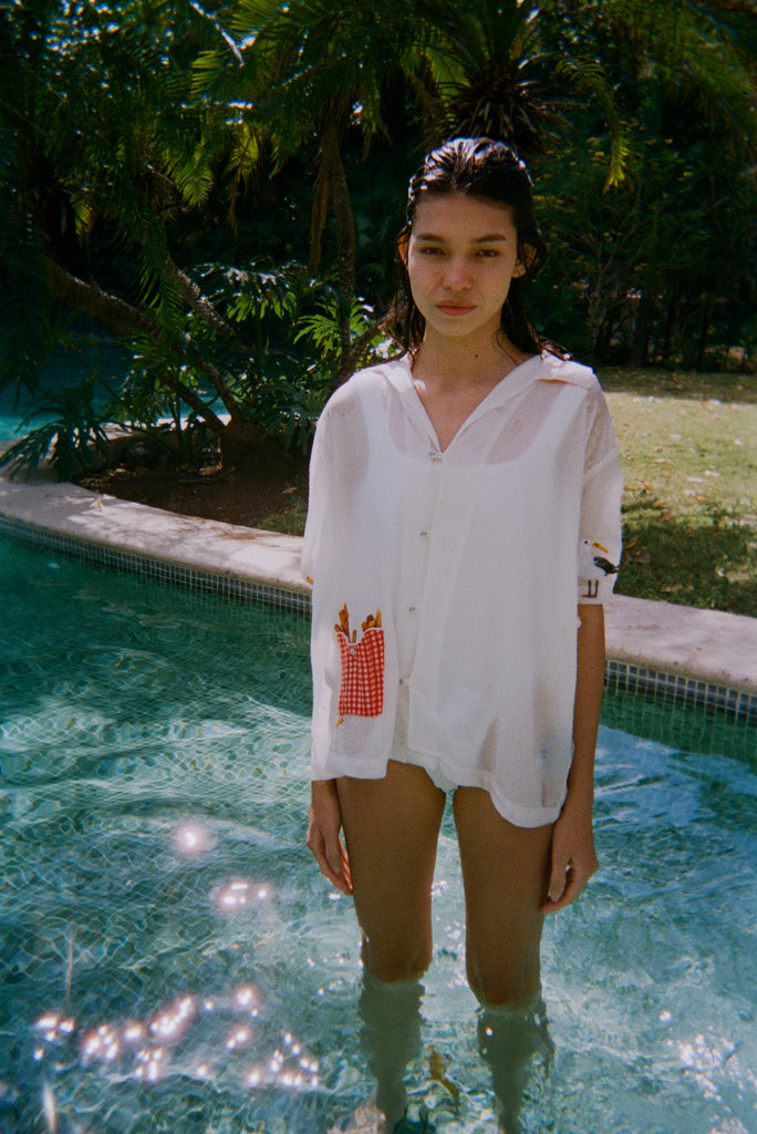Woman standing in a pool wearing a sheer white short-sleeve shirt with a red gingham pocket embroidered with French fries and a seagull embroidered on the sleeve.