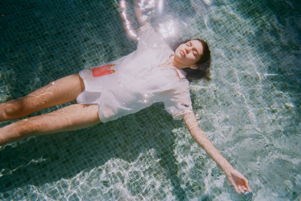  Woman floating in a pool wearing a white button-up shirt with a red gingham pocket embroidered with French fries and a seagull motif on the sleeve.