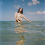 A woman stands waist-deep in clear ocean water, wearing the 'Starfish Strut' Hand Towel Top.  Her eyes are closed, head tilted gently, and her reflection shimmers in the water below. 