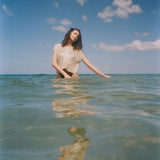 A woman stands waist-deep in clear ocean water, wearing the 'Starfish Strut' Hand Towel Top.  Her eyes are closed, head tilted gently, and her reflection shimmers in the water below. 