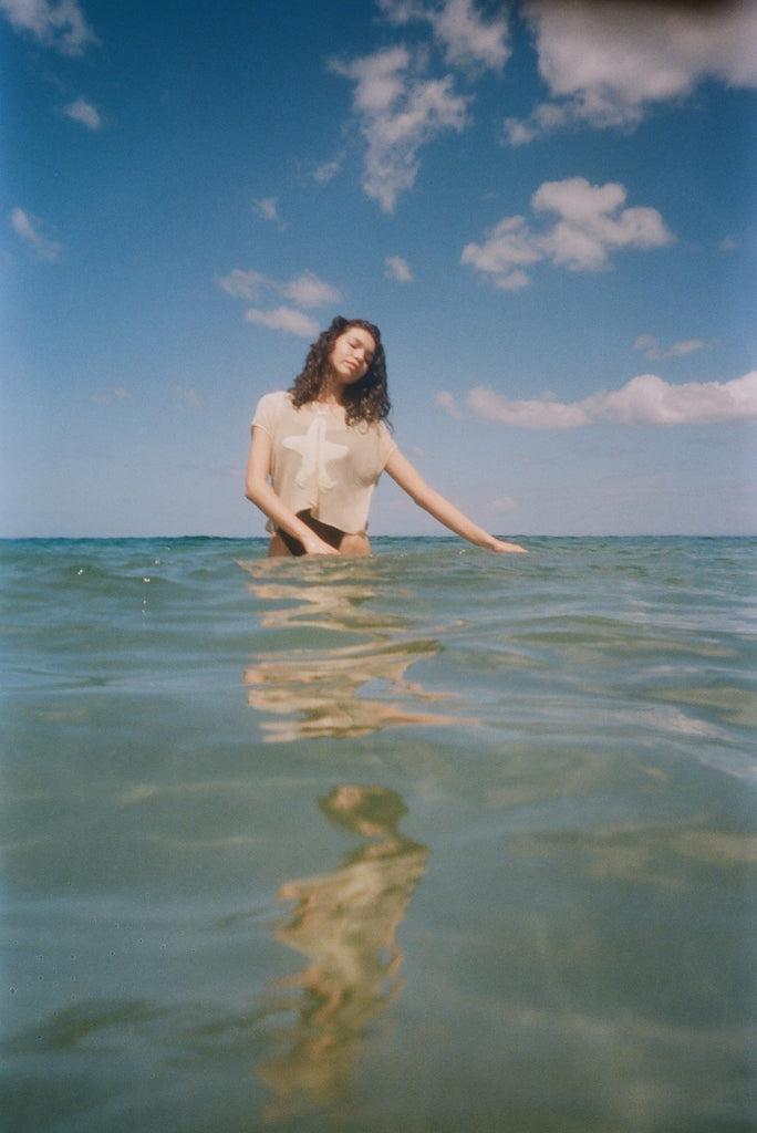A woman stands waist-deep in clear ocean water, wearing the 'Starfish Strut' Hand Towel Top.  Her eyes are closed, head tilted gently, and her reflection shimmers in the water below. 