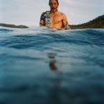 A man with wet hair and a mustache stands chest-deep in the ocean, wearing a partially open 'Perfect Cast' Shirt. The foreground is dominated by rippling dark blue water.