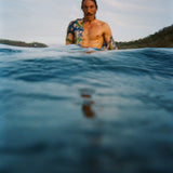 A man with wet hair and a mustache stands chest-deep in the ocean, wearing a partially open 'Perfect Cast' Shirt. The foreground is dominated by rippling dark blue water.