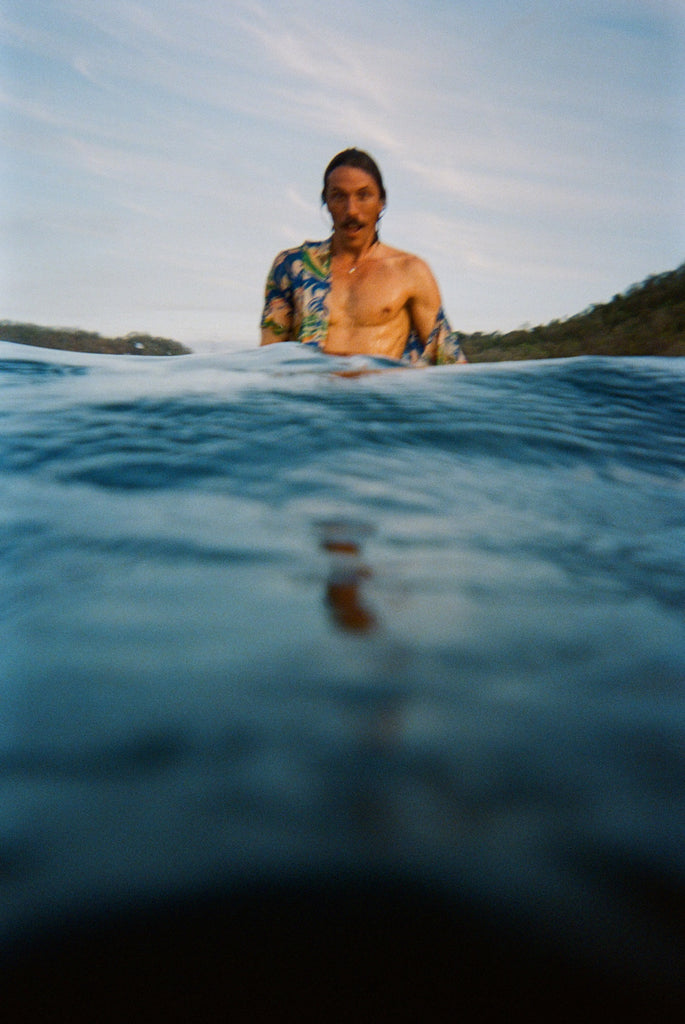 A man with wet hair and a mustache stands chest-deep in the ocean, wearing a partially open 'Perfect Cast' Shirt. The foreground is dominated by rippling dark blue water.