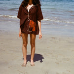 Cropped image of female model wearing the brown button-down 'Bisque Averse' Cabana and standing facing the camera on a sunny beach.