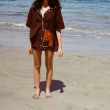Cropped image of female model wearing the brown button-down 'Bisque Averse' Cabana and standing facing the camera on a sunny beach.