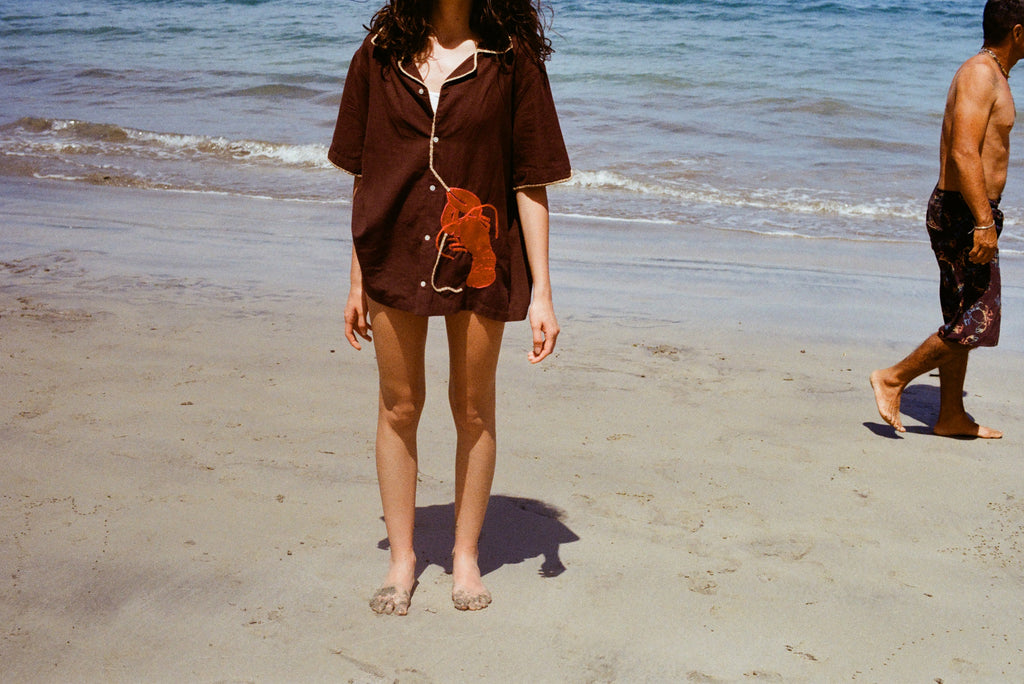 Cropped image of female model wearing the brown button-down 'Bisque Averse' Cabana and standing facing the camera on a sunny beach.
