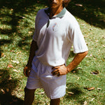 A man wearing a white terry cloth polo shirt with a green collar and matching green accents. He is also wearing white drawstring shorts, with one hand on his hip.