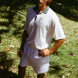 A man wearing a white terry cloth polo shirt with a green collar and matching green accents. He is also wearing white drawstring shorts, with one hand on his hip.