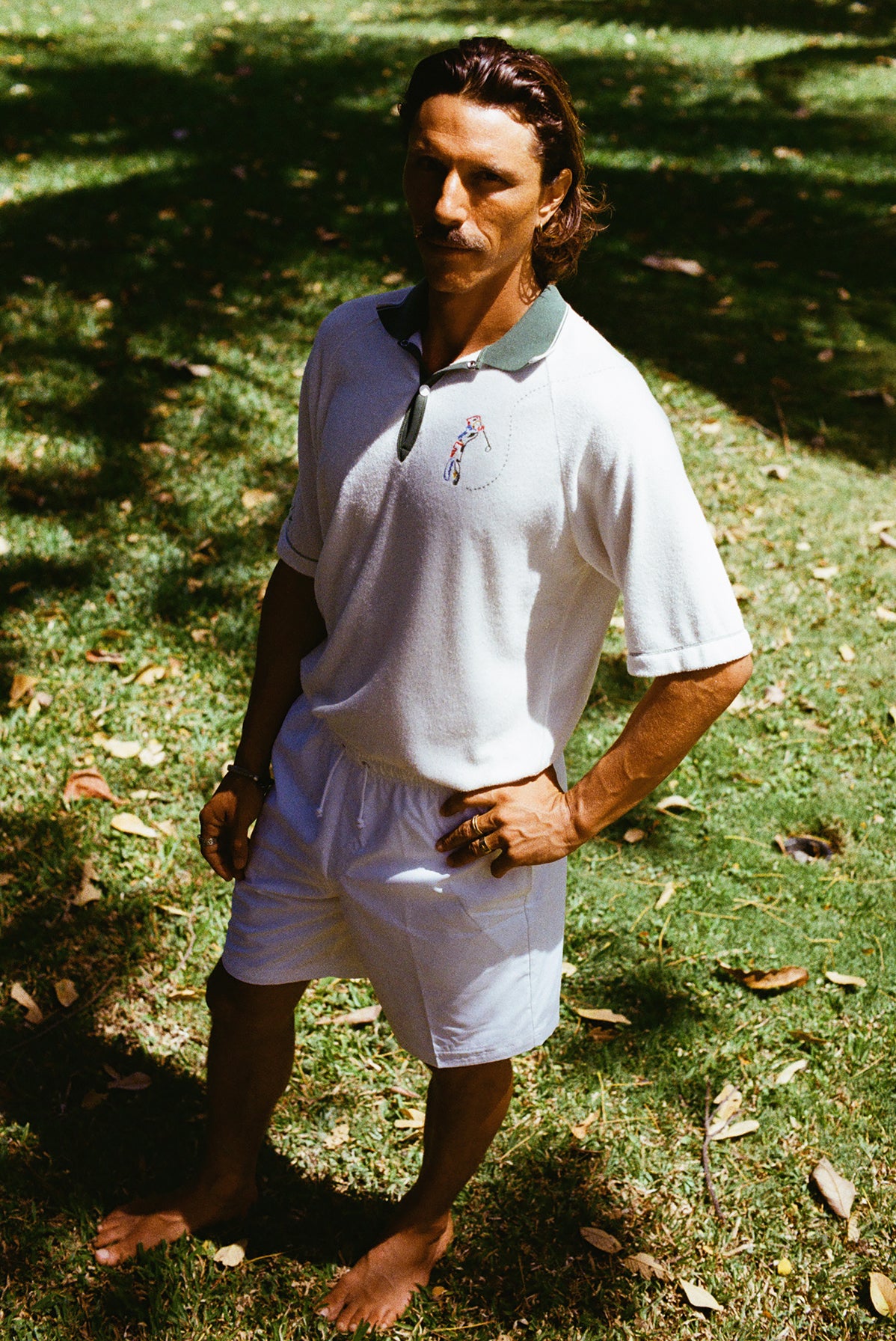 A man wearing a white terry cloth polo shirt with a green collar and matching green accents. He is also wearing white drawstring shorts, with one hand on his hip.