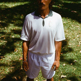A man wearing a white terry cloth polo shirt with a green collar and matching green accents. He is also wearing white drawstring shorts.