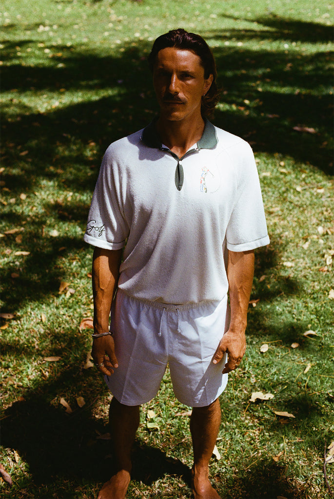 A man wearing a white terry cloth polo shirt with a green collar and matching green accents. He is also wearing white drawstring shorts.