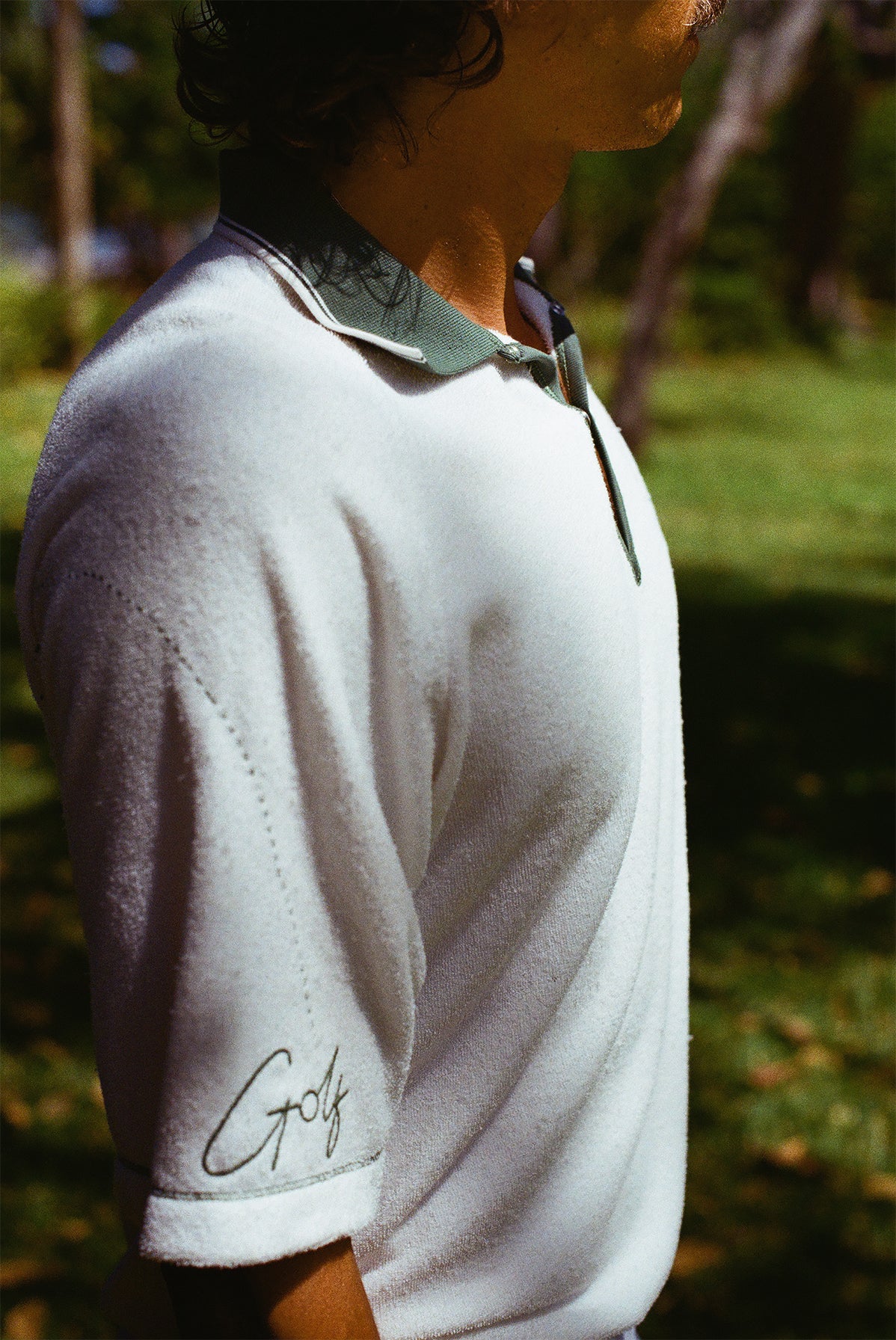 Close-up side view of a man wearing a white terry cloth polo shirt with a green collar and placket. The left sleeve features green script embroidery reading 'Golf' with a dotted line extending upward toward the shoulder. 