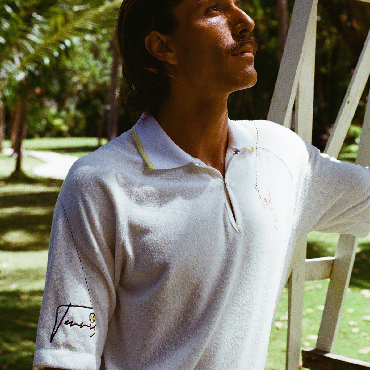 Man wearing a white terry cloth polo with embroidered tennis graphics on the chest and sleeve, standing beside a vintage wooden umpire chair.
