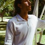 Man wearing a white terry cloth polo with embroidered tennis graphics on the chest and sleeve, standing beside a vintage wooden umpire chair.