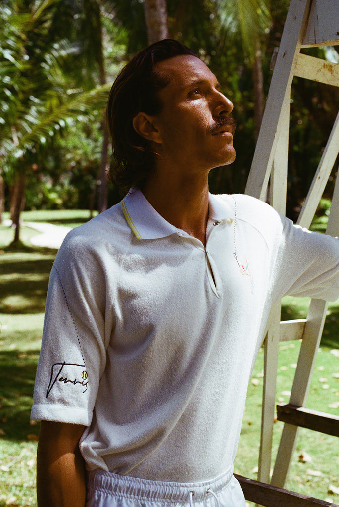 Man wearing a white terry cloth polo with embroidered tennis graphics on the chest and sleeve, standing beside a vintage wooden umpire chair.