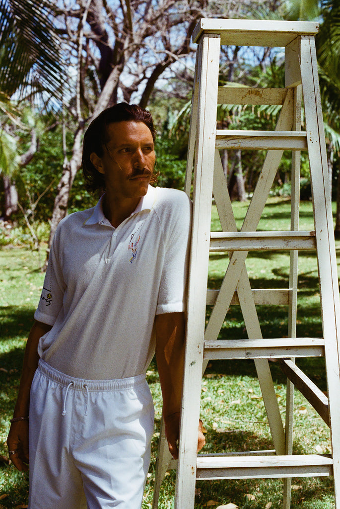 Man wearing a white terry cloth polo with embroidered tennis graphics on the chest and sleeve.