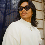 man with shoulder length black hair looking to the left; wearing beige sweater and black thick framed sunglasses