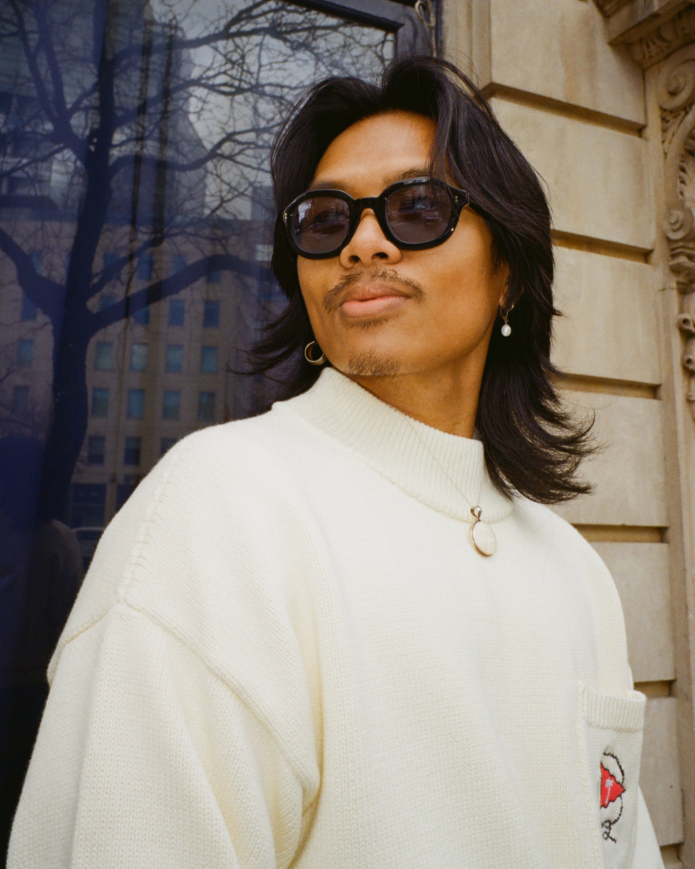 man with shoulder length black hair looking to the left; wearing beige sweater and black thick framed sunglasses