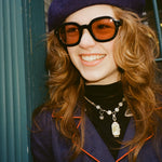 Young female model with long reddish brown hair wearing Lesca Lunetier x Tombolo Lenses in Coral  while leaning against a forest green pillar with a navy beret and navy buttoned coat that has a red outlined lapel.