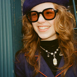 Young female model with long reddish brown hair wearing Lesca Lunetier x Tombolo Lenses in Coral  while leaning against a forest green pillar with a navy beret and navy buttoned coat that has a red outlined lapel.