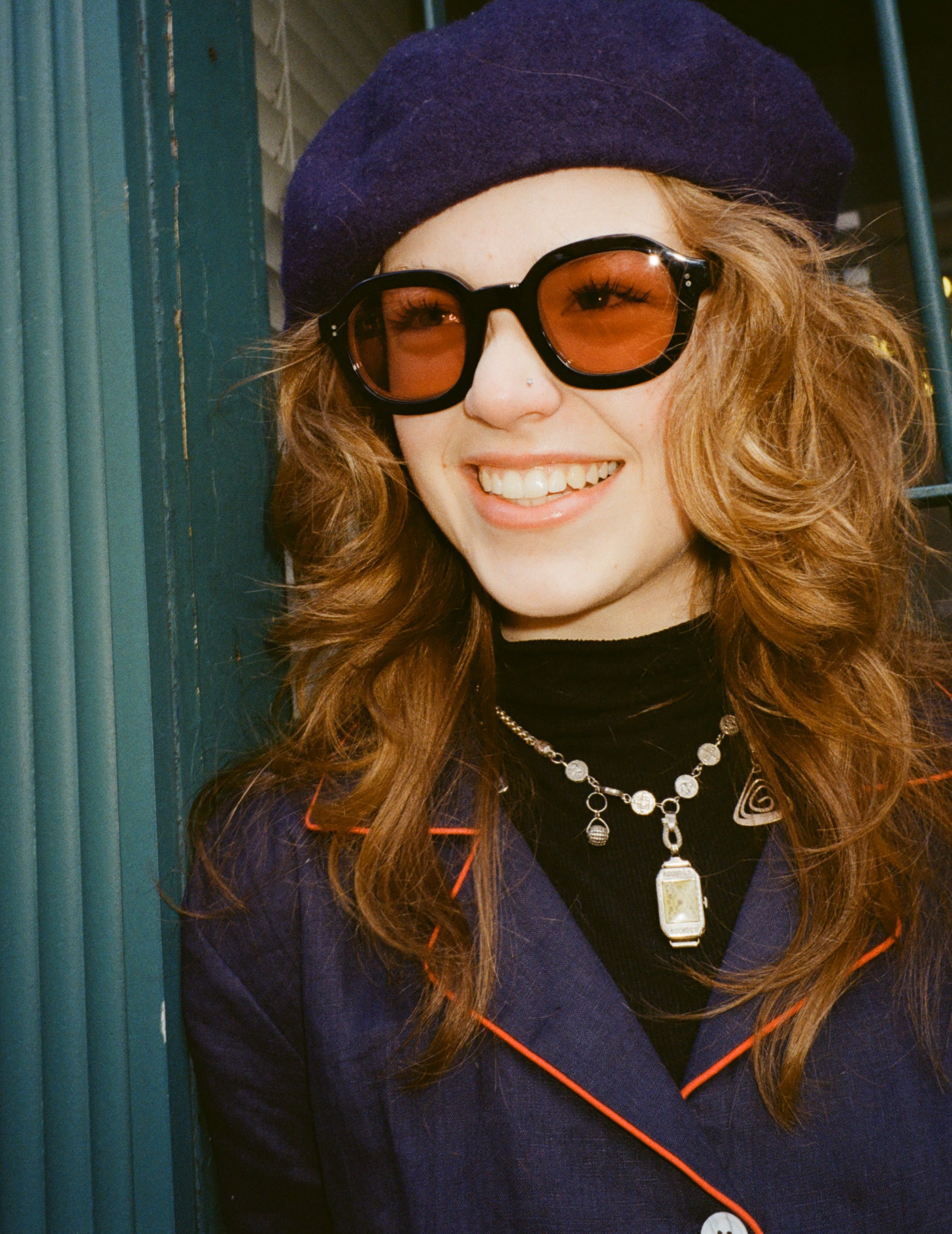 Young female model with long reddish brown hair wearing Lesca Lunetier x Tombolo Lenses in Coral  while leaning against a forest green pillar with a navy beret and navy buttoned coat that has a red outlined lapel.
