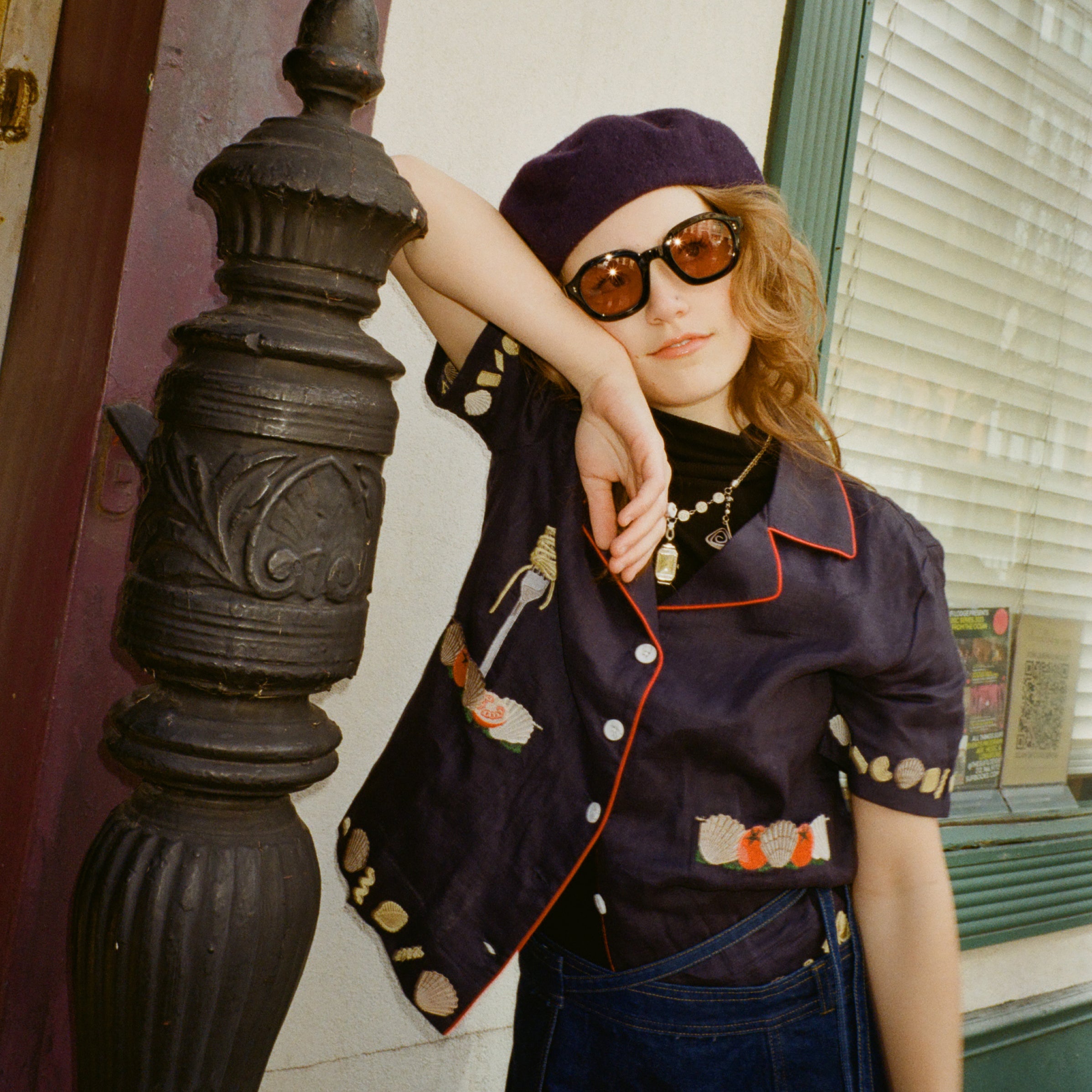 Young female model wearing Lesca Lunetier x Tombolo lenses in coral leaning against a pillar wearing a navy beret and Tombolo "Vongole" Cabana in Navy.