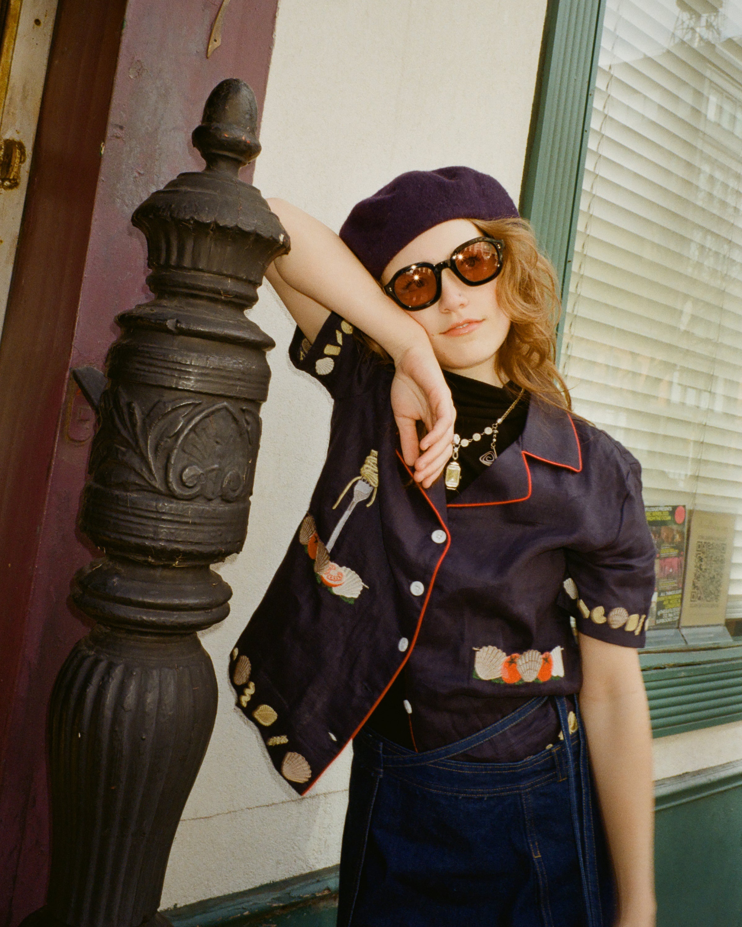 Young female model wearing Lesca Lunetier x Tombolo lenses in coral leaning against a pillar wearing a navy beret and Tombolo "Vongole" Cabana in Navy.