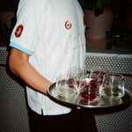 A person wearing a white soccer jersey with light blue trim and gold script lettering, holding a silver tray with several clear drinking glasses in a dimly lit indoor setting.