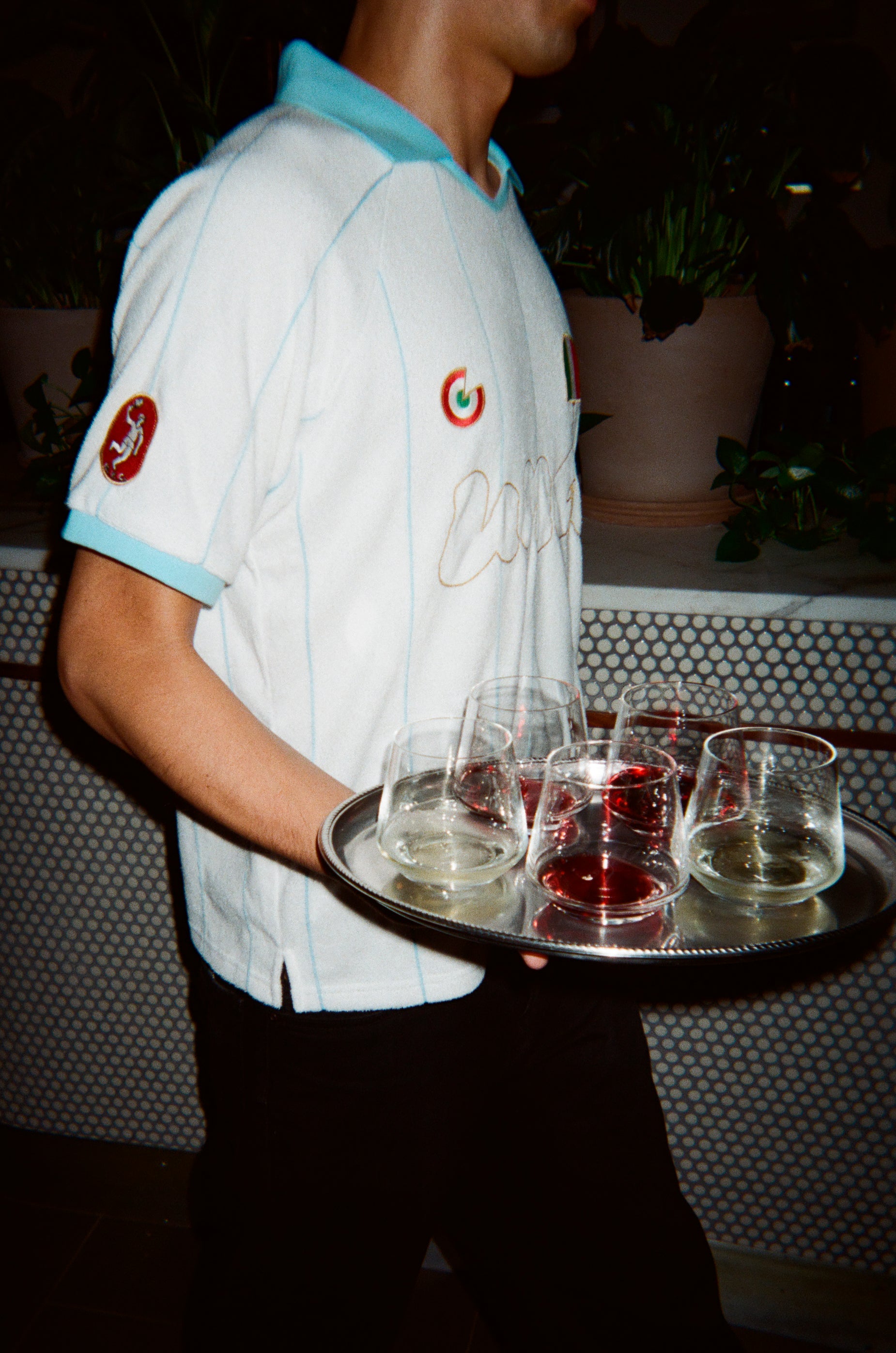 A person wearing a white soccer jersey with light blue trim and gold script lettering, holding a silver tray with several clear drinking glasses in a dimly lit indoor setting.