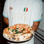 A person wearing a white soccer-style jersey with light blue trim and gold script lettering holds a freshly baked pizza on a white plate. The jersey features a small Italian flag graphic and a circular logo on the chest.