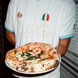 A person wearing a white soccer-style jersey with light blue trim and gold script lettering holds a freshly baked pizza on a white plate. The jersey features a small Italian flag graphic and a circular logo on the chest.