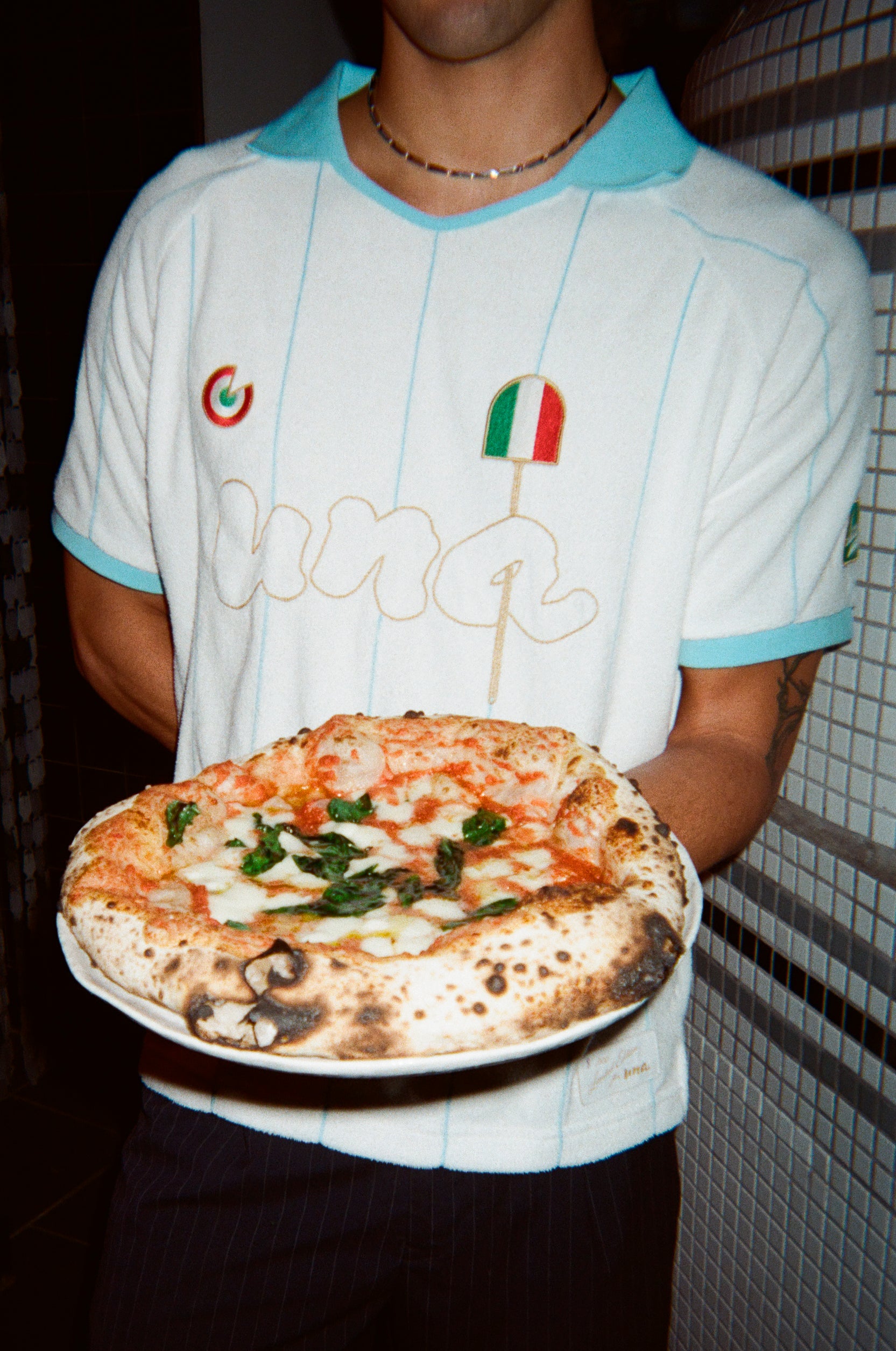 A person wearing a white soccer-style jersey with light blue trim and gold script lettering holds a freshly baked pizza on a white plate. The jersey features a small Italian flag graphic and a circular logo on the chest.