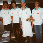 Four people wearing matching white and light blue Italian-themed sports jerseys, with one holding a pizza in a restaurant setting.