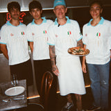 Four people wearing matching white and light blue Italian-themed sports jerseys, with one holding a pizza in a restaurant setting.