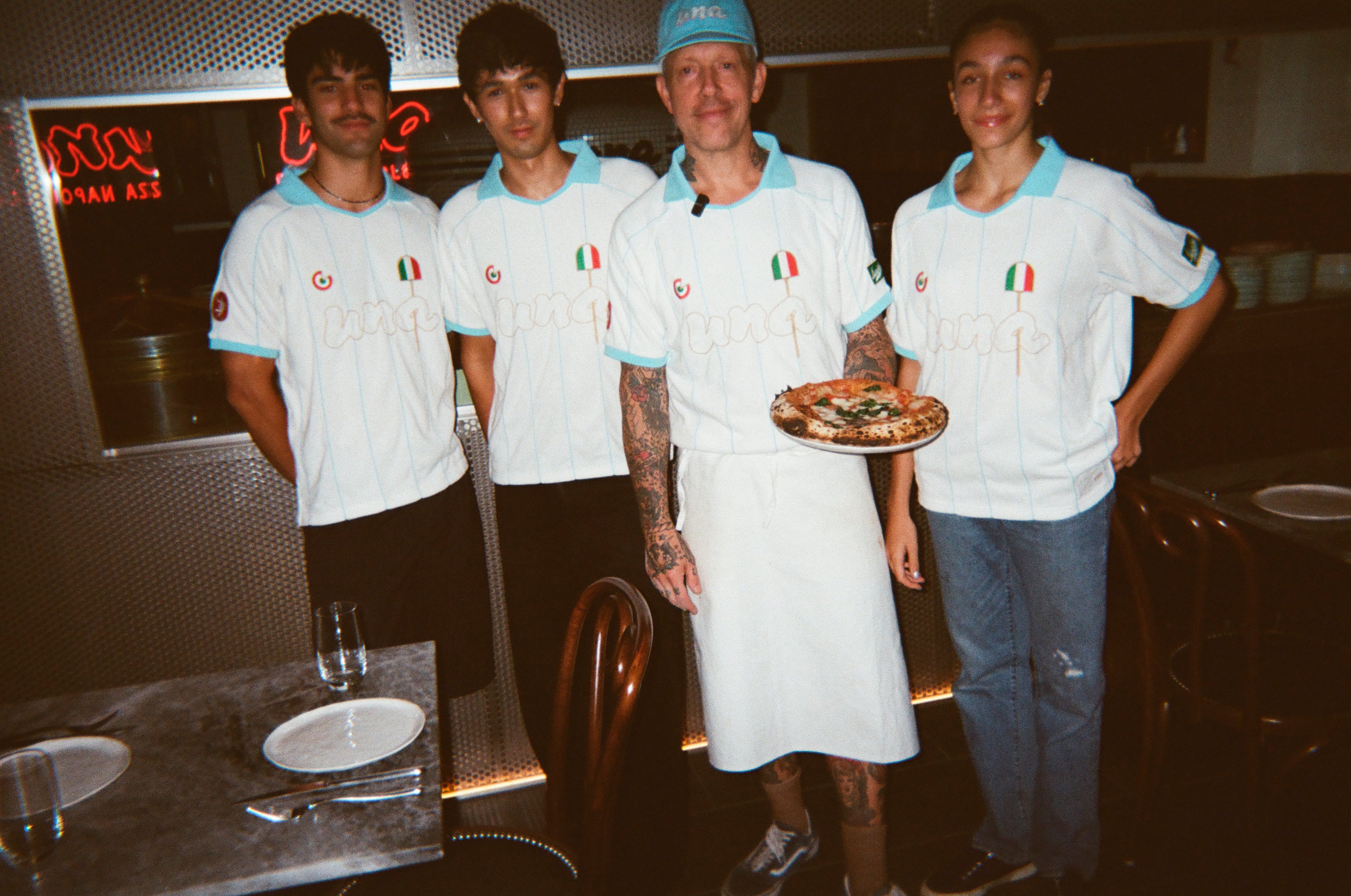 Four people wearing matching white and light blue Italian-themed sports jerseys, with one holding a pizza in a restaurant setting.