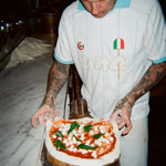 A tattooed pizza chef wearing a white and light blue striped polo shirt with a small Italian flag logo, holding a freshly baked pizza on a metal tray in a dimly lit kitchen.