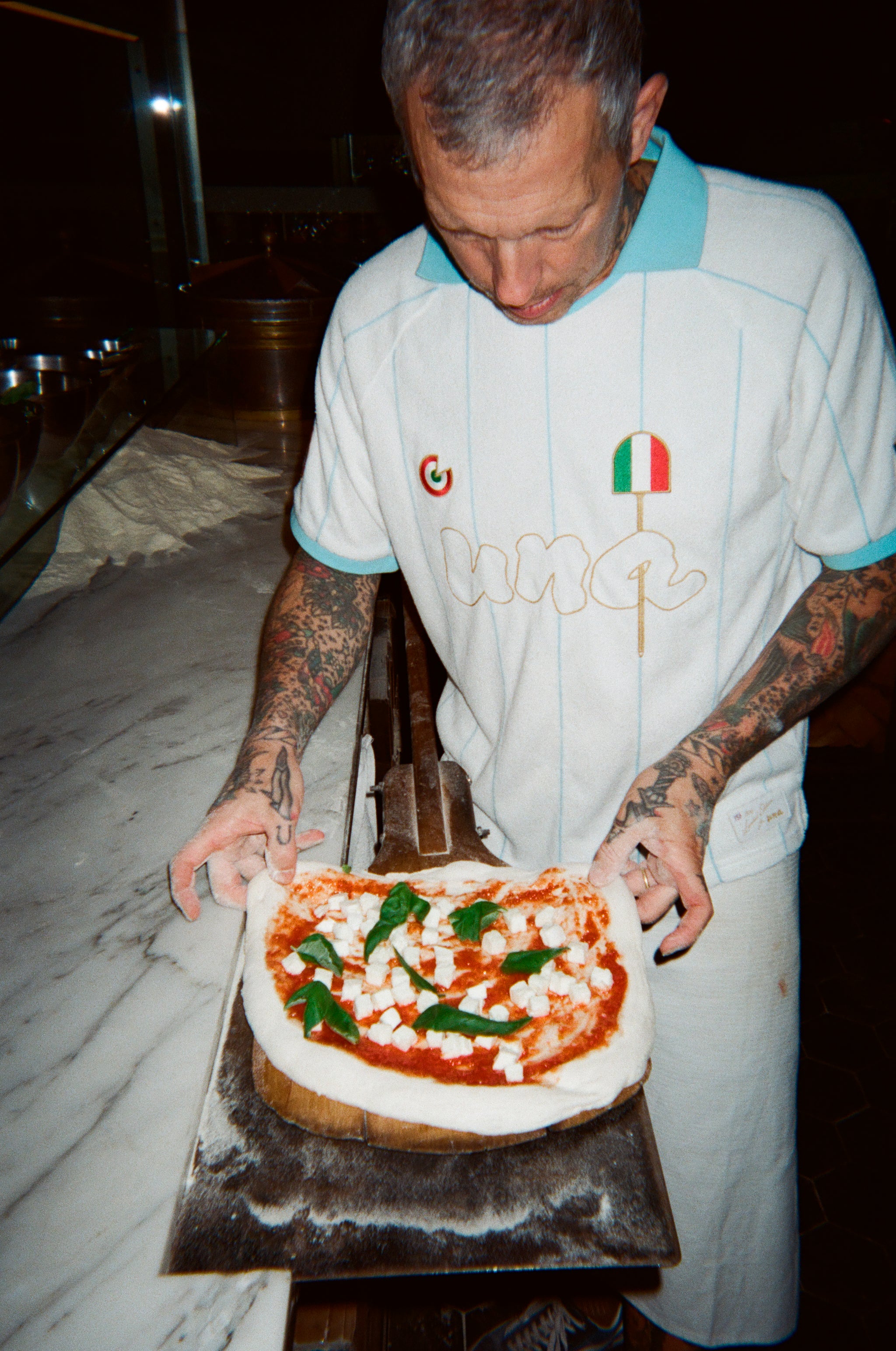 A tattooed pizza chef wearing a white and light blue striped polo shirt with a small Italian flag logo, holding a freshly baked pizza on a metal tray in a dimly lit kitchen.