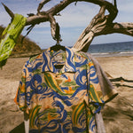 A shot of our 'Perfect Cast' Hawaiian Shirt hanging on a tree branch at the beach. A few Tombolo shirts hang behind it. 