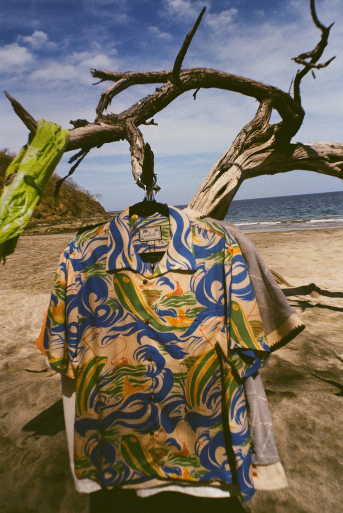 A shot of our 'Perfect Cast' Hawaiian Shirt hanging on a tree branch at the beach. A few Tombolo shirts hang behind it. 