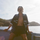 Brunette man wearing black short-sleeve terrycloth surf cabana, fully unzipped while sitting on the edge of a boat.