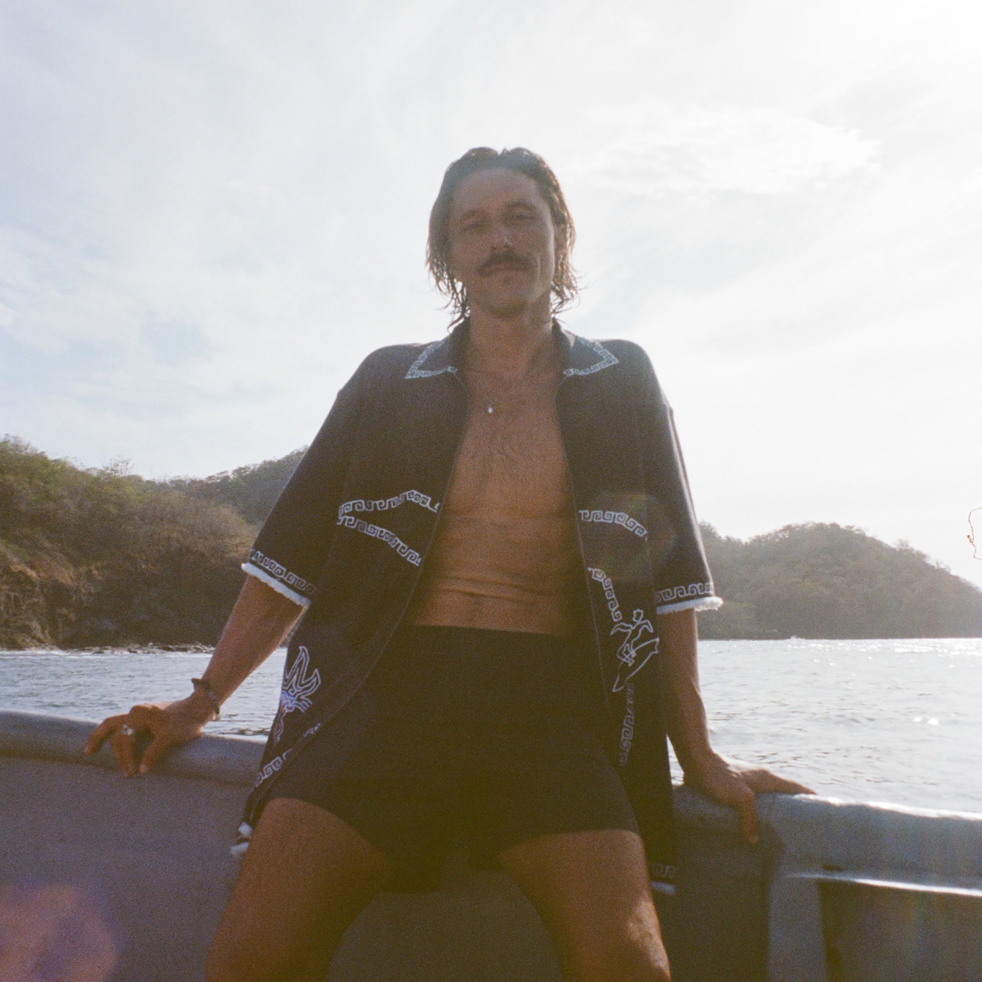 Brunette man wearing black short-sleeve terrycloth surf cabana, fully unzipped while sitting on the edge of a boat.