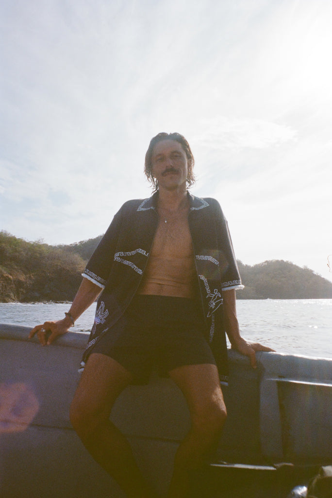 Brunette man wearing black short-sleeve terrycloth surf cabana, fully unzipped while sitting on the edge of a boat.