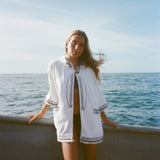Blonde woman standing on a boat facing the camera, wearing white hooded terry cloth beach towel top with black embroidered trim, over a black bikini. 