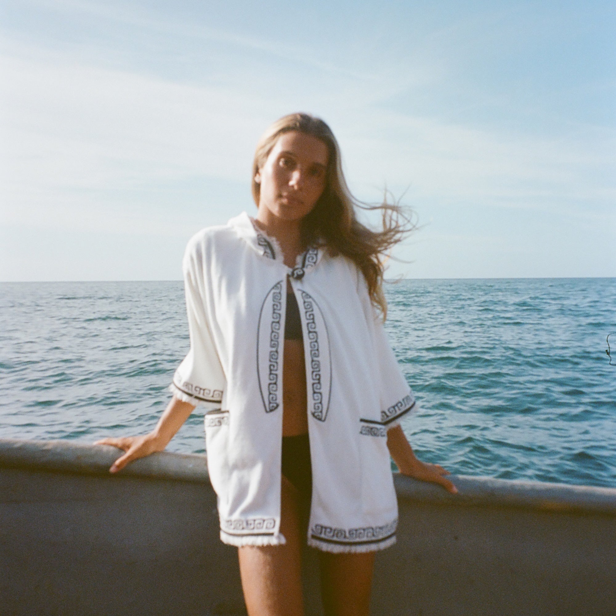 Blonde woman standing on a boat facing the camera, wearing white hooded terry cloth beach towel top with black embroidered trim, over a black bikini. 